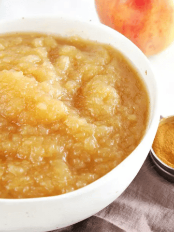 Pressure Cooker Applesauce in a white bowl, a whole apple and a small container of cinnamon on the side