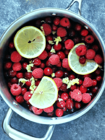 overhead view of Ginger Cranberry Sauce being made: large stainless saucepan filled with whole fresh cranberries, raspberries, lemon slices, and finely minced ginger.