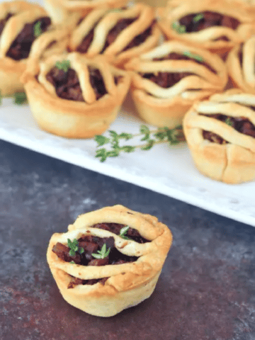 Mini vegan sausage pies on a white square platter dressed with sprigs of fresh thyme, with one mini pie off the plate in front.