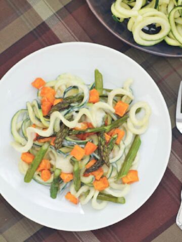 overhead view of roasted sweet potato asparagus pasta using spiralized zucchini as noodles, on a white plate with a brown plaid tabletop, a side of zucchini spirals next to dish.