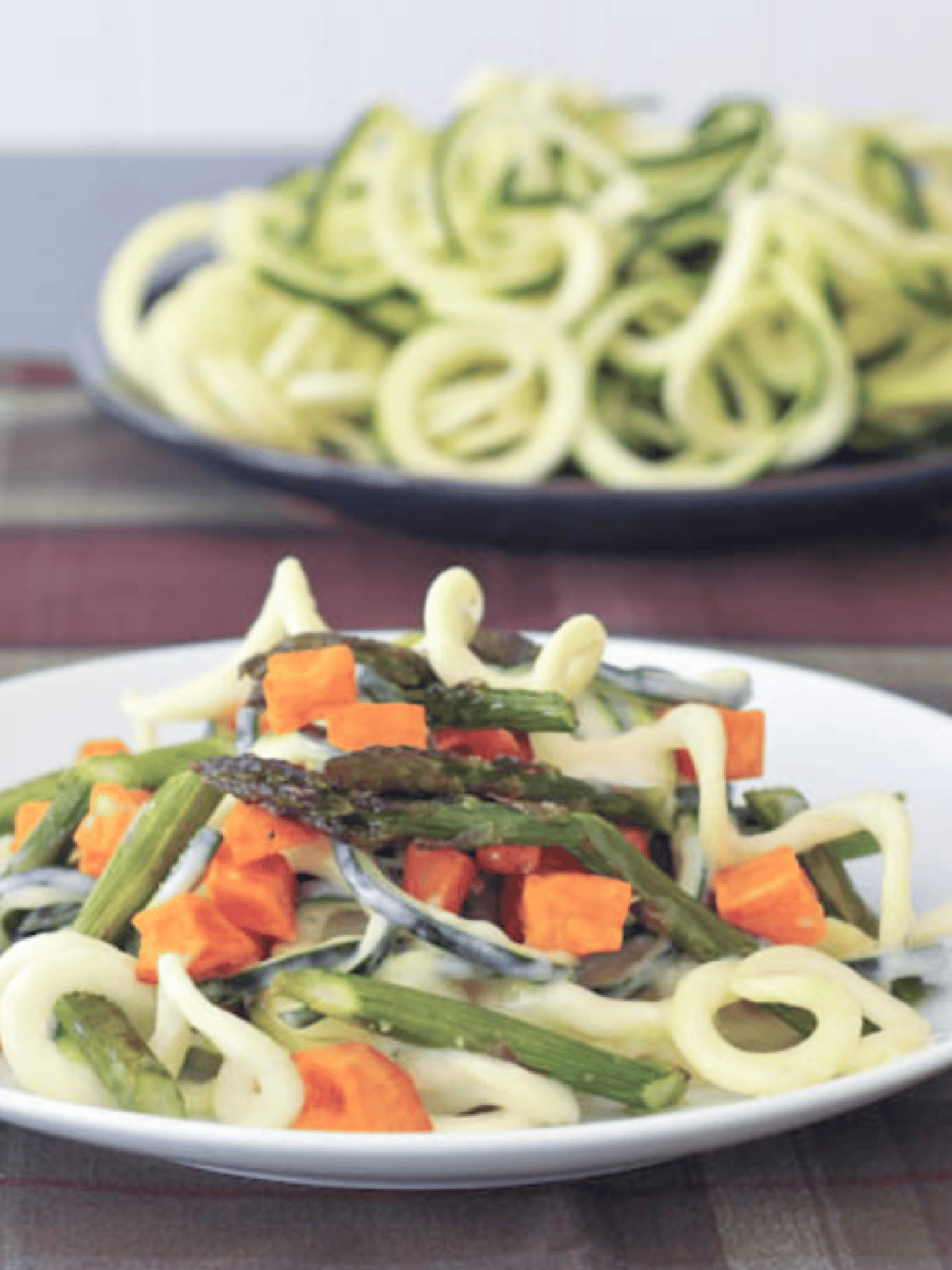 roasted sweet potato asparagus pasta using spiralized zucchini as noodles, on a white plate with a brown plaid tabletop, a side of zucchini spirals next to dish.