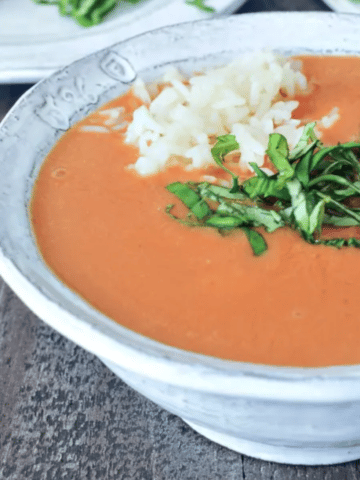 Tomato orange soup in a rustic white bowl, garnished with chopped basil and white rice, a plate with crusty baguette on the side.