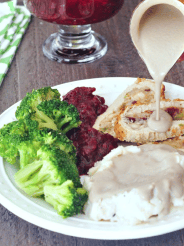 vegan mushroom gravy being poured over mashed potatoes and a vegan roast, broccoli and cranberry sauce also on the plate.