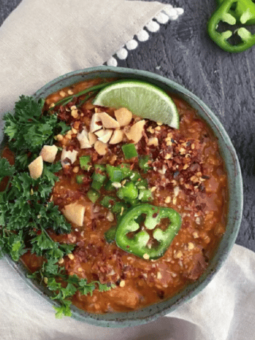 overhead view of bright orange spicy vegan peanut soup in a rustic bowl, topped with parsley, sliced jalapeno, chopped peanuts.