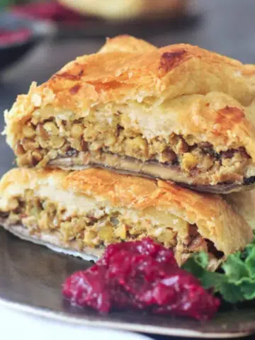 Individual portobello wellington cut in half and stacked on a dark grey plate to show inside filling of lentil veggie stuffing, kale salad and bright red cranberry sauce on the side