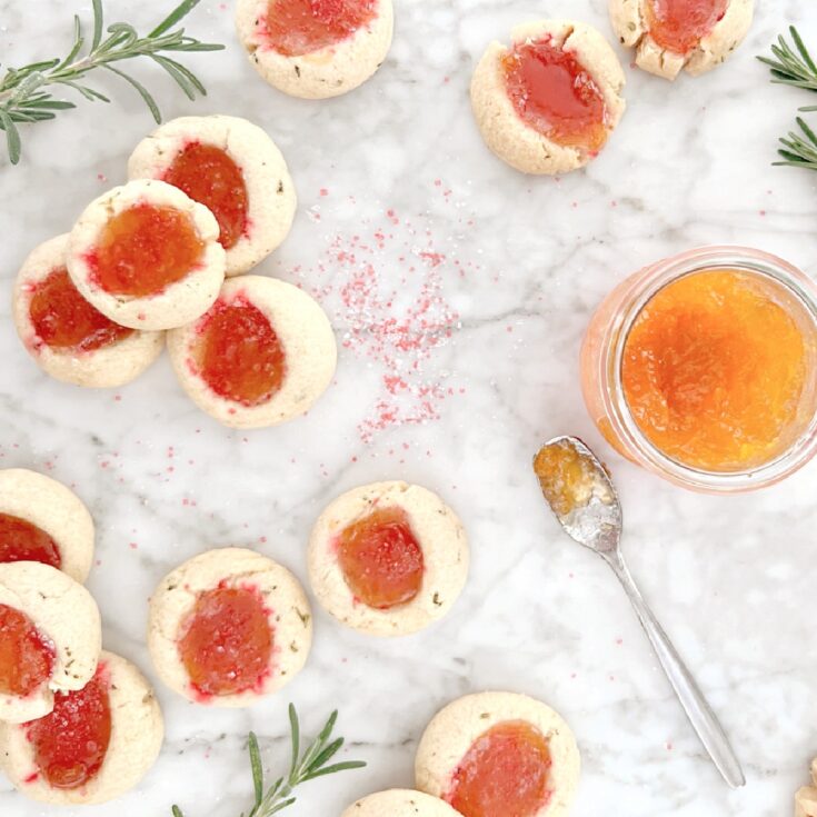 overhead view of sparkly orange apricot jam thumbprint cookies scattered across a white marble table with a jar of apricot jam, a spoon with jam on it, sprigs of fresh rosemary (which is in the cookie), and sparkly sanding sugar sprinkled around.