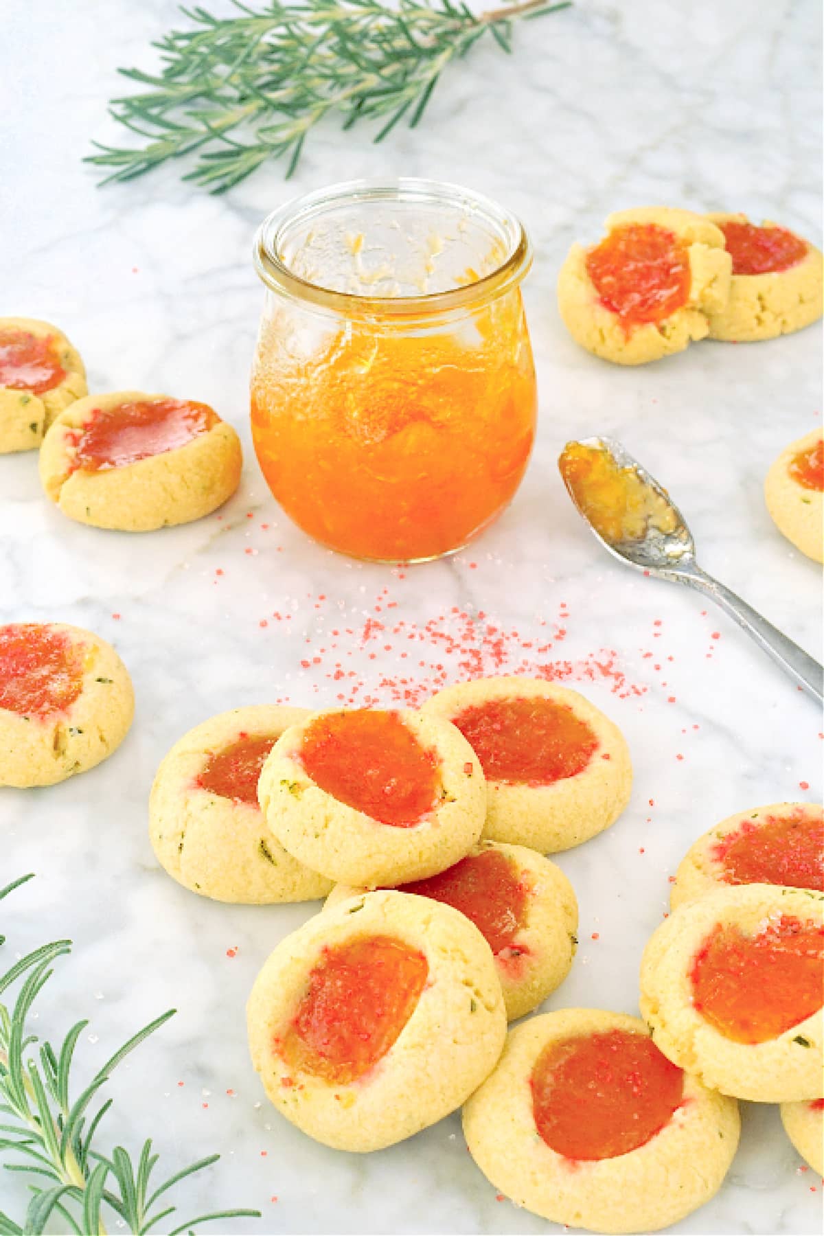 sparkly orange apricot jam thumbprint cookies scattered across a white marble table with a jar of apricot jam, a spoon with jam on it, sprigs of fresh rosemary (which is in the cookie), and sparkly sanding sugar sprinkled around.
