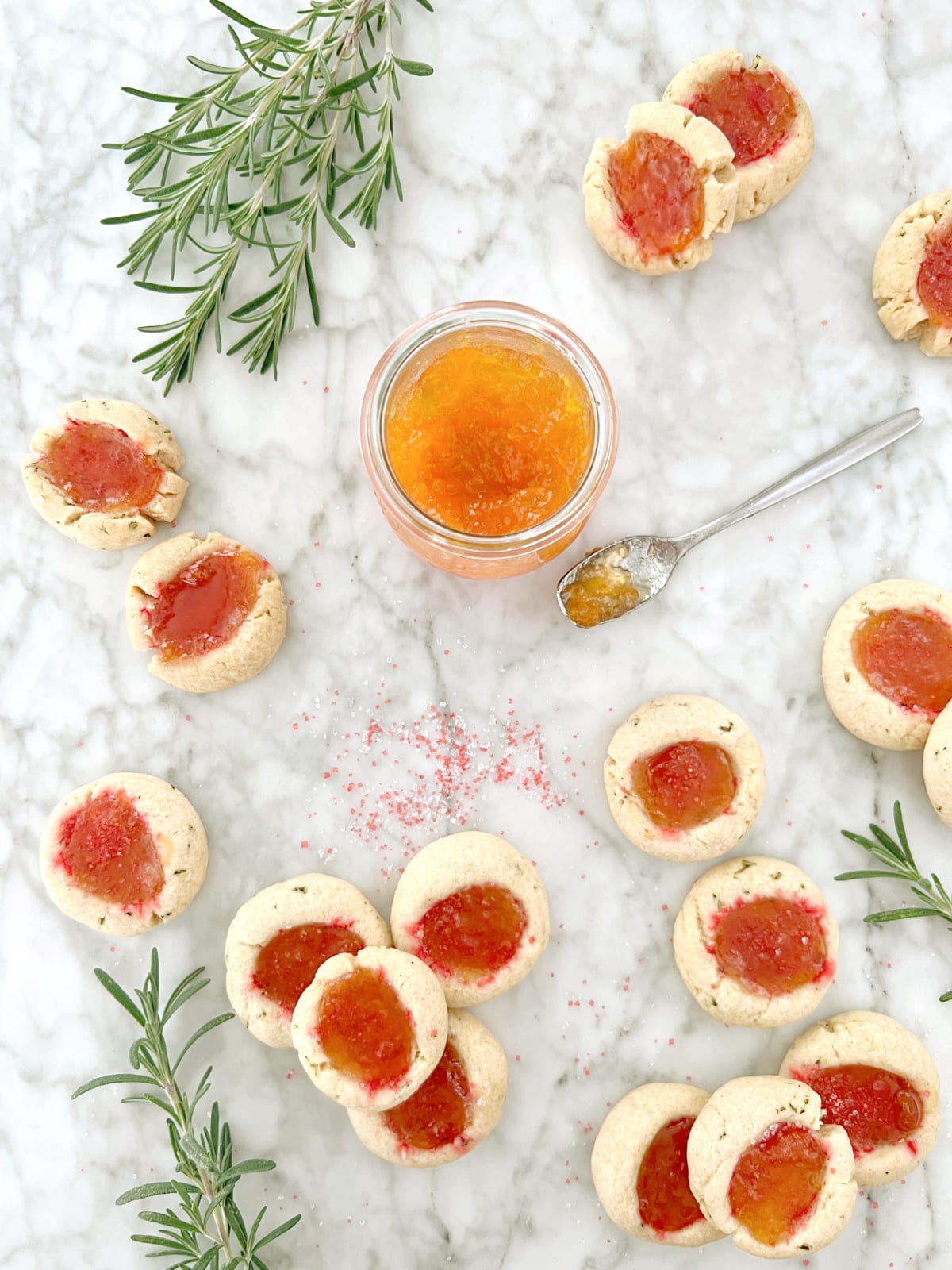 overhead view of sparkly orange apricot jam thumbprint cookies scattered across a white marble table with a jar of apricot jam, a spoon with jam on it, sprigs of fresh rosemary (which is in the cookie), and sparkly sanding sugar sprinkled around.
