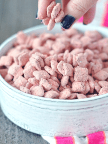 hand reaching in to bowl of Powder Pink Strawberry Puppy Chow (a strawberry powder and chocolate coated cereal treat)