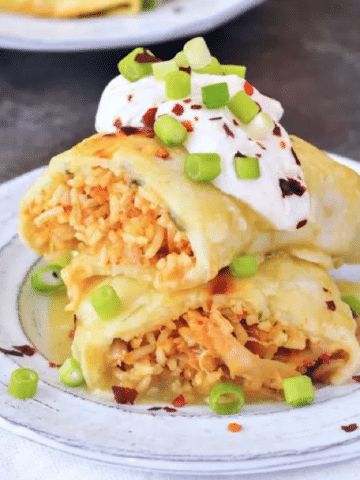 Vegan Buffalo Jackfruit Enchiladas sliced in half and stacked on a grey rustic plate, insides filled with orange buffalo sauced, shredded jackfruit, rice, topped with sour cream and sliced green onion.