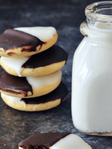 stack of four black and white cookies (a cakey cookie with half vanilla frosting, half chocolate frosting) next to a small glass bottle of milk.