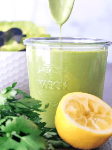 a jar of Green Goddess dressing with a spoon coming out of it, a squeezed lemon and a bunch of herbs on the side.