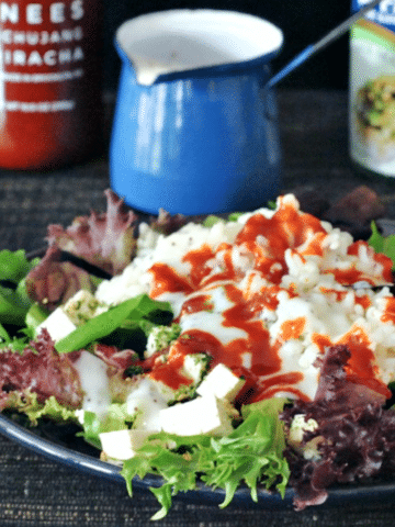 a green salad with diced tofu, sriracha, sticky rice and furikake flakes on a dark blue dish, drizzled with poppyseed dressing. a bottle of sriracha and jar of furikake in background.