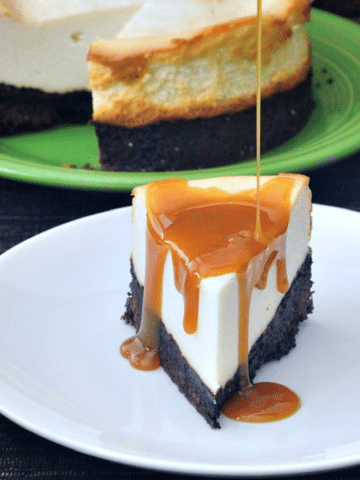 Brownie Bottomed Cheesecake slice on plate with caramel sauce poured over, whole cheesecake in background