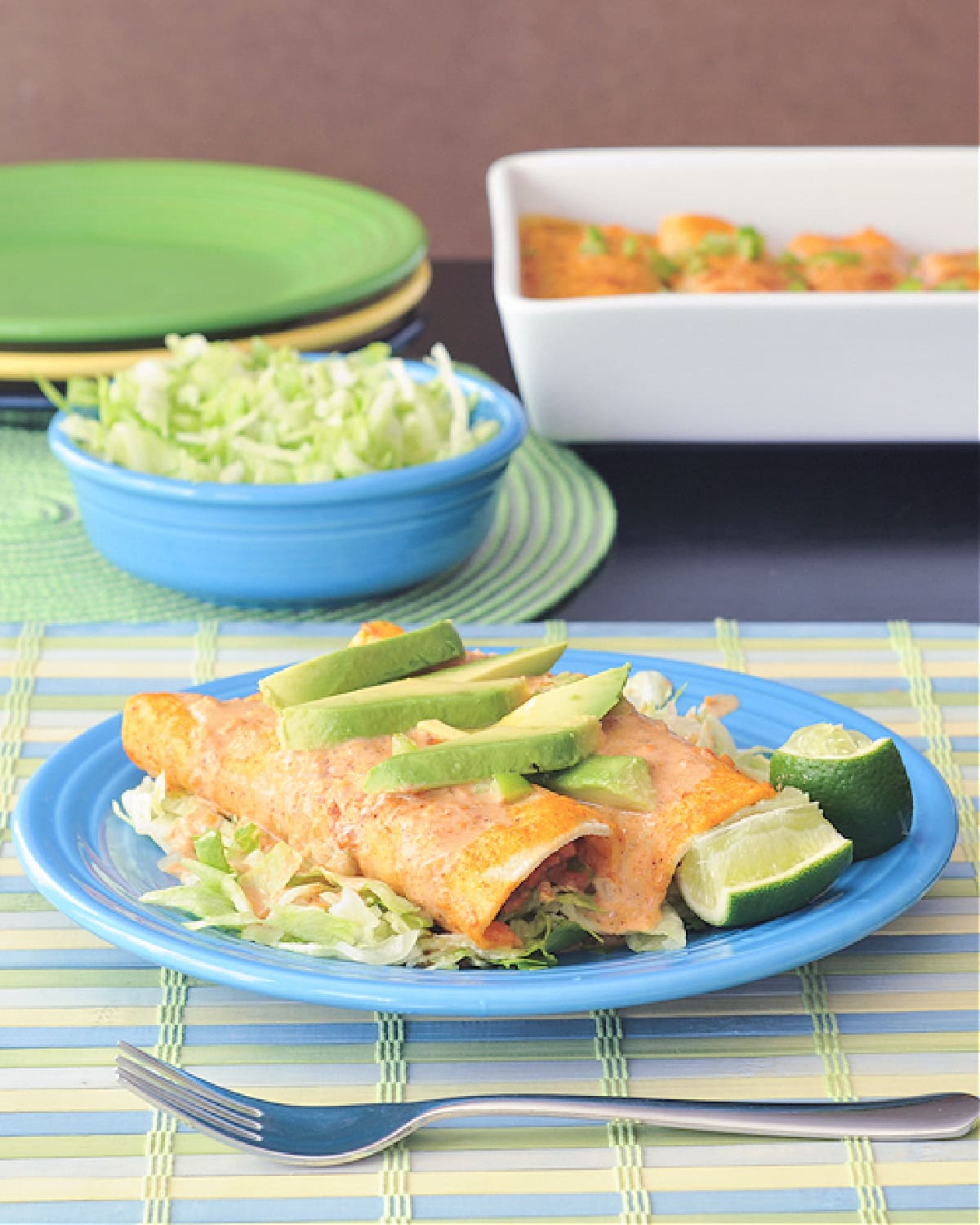 two tortilla soup enchiladas on a blue Fiestaware plate: creamy orange colored sauce tops bean, corn, pepper and rice stuffed enchiladas, topped with sliced avocado. two lime wedges on the side. blue bowl of shredded lettuce and white ceramic baking dish full of enchiladas in background.