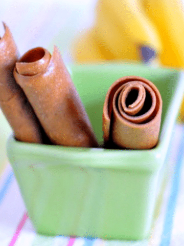 three peanut butter banana fruit roll (fruit leather made from pureed peanut butter and bananas) rolled up in tubes and sticking out of a small green ceramic loaf pan. a bunch of fresh bananas blurred in background, surface is a pastel striped napkin.