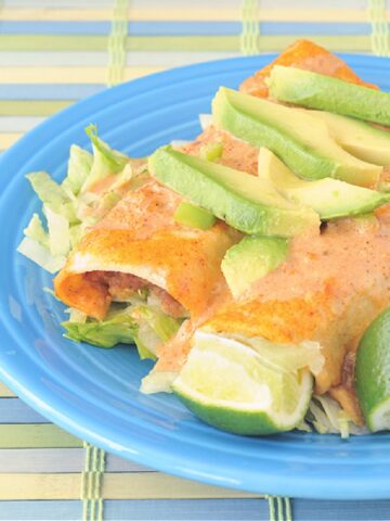 two tortilla soup enchiladas on a blue Fiestaware plate: creamy orange colored sauce tops bean, corn, pepper and rice stuffed enchiladas, topped with sliced avocado. two lime wedges on the side.