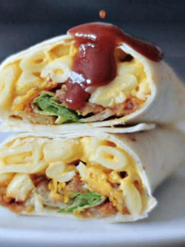 A BBQ mac and cheese burrito sliced in half on a white plate, with BBQ sauce dripping on top.