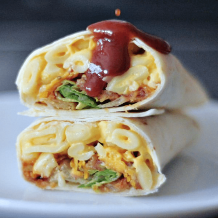 A BBQ mac and cheese burrito sliced in half on a white plate, with BBQ sauce dripping on top.