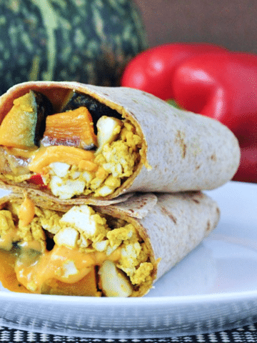 One cheesy kabocha breakfast burrito on a white plate, sliced in half to show filling (kabocha squash, vegan eggs, melty dairy free cheese).
