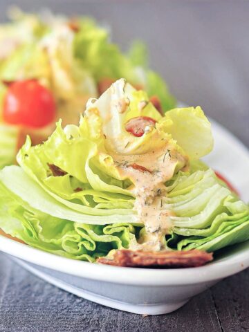 close up of one mini wedge salad on a long white oval platter, dressed with sliced grape tomatoes, vegan bacon pieces, and chipotle ranch dressing. more mini wedge salads on platter are blurred in background.