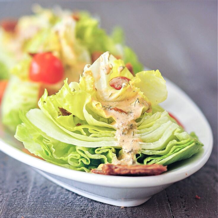 close up of one mini wedge salad on a long white oval platter, dressed with sliced grape tomatoes, vegan bacon pieces, and chipotle ranch dressing. more mini wedge salads on platter are blurred in background.