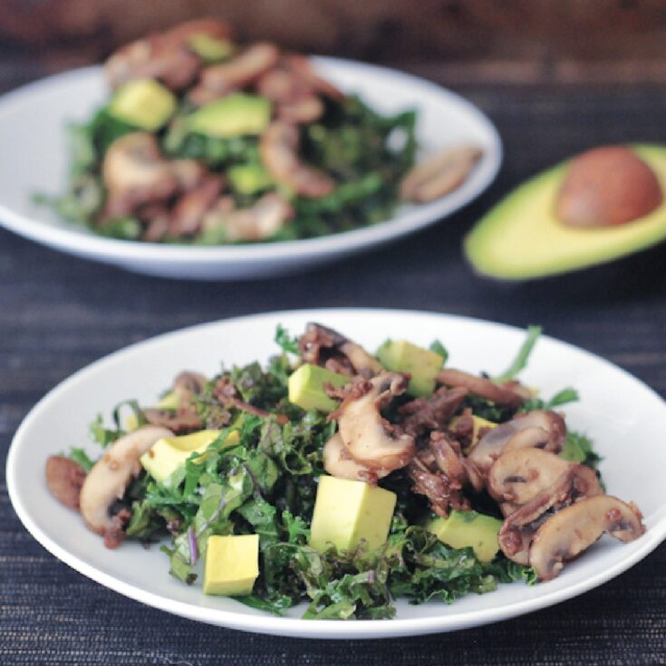 two white plates of kale salad with warm bacon dressing, one plate blurred in background with a half avocado. salad includes kale, cubed avocado, bacon flavored mushrooms