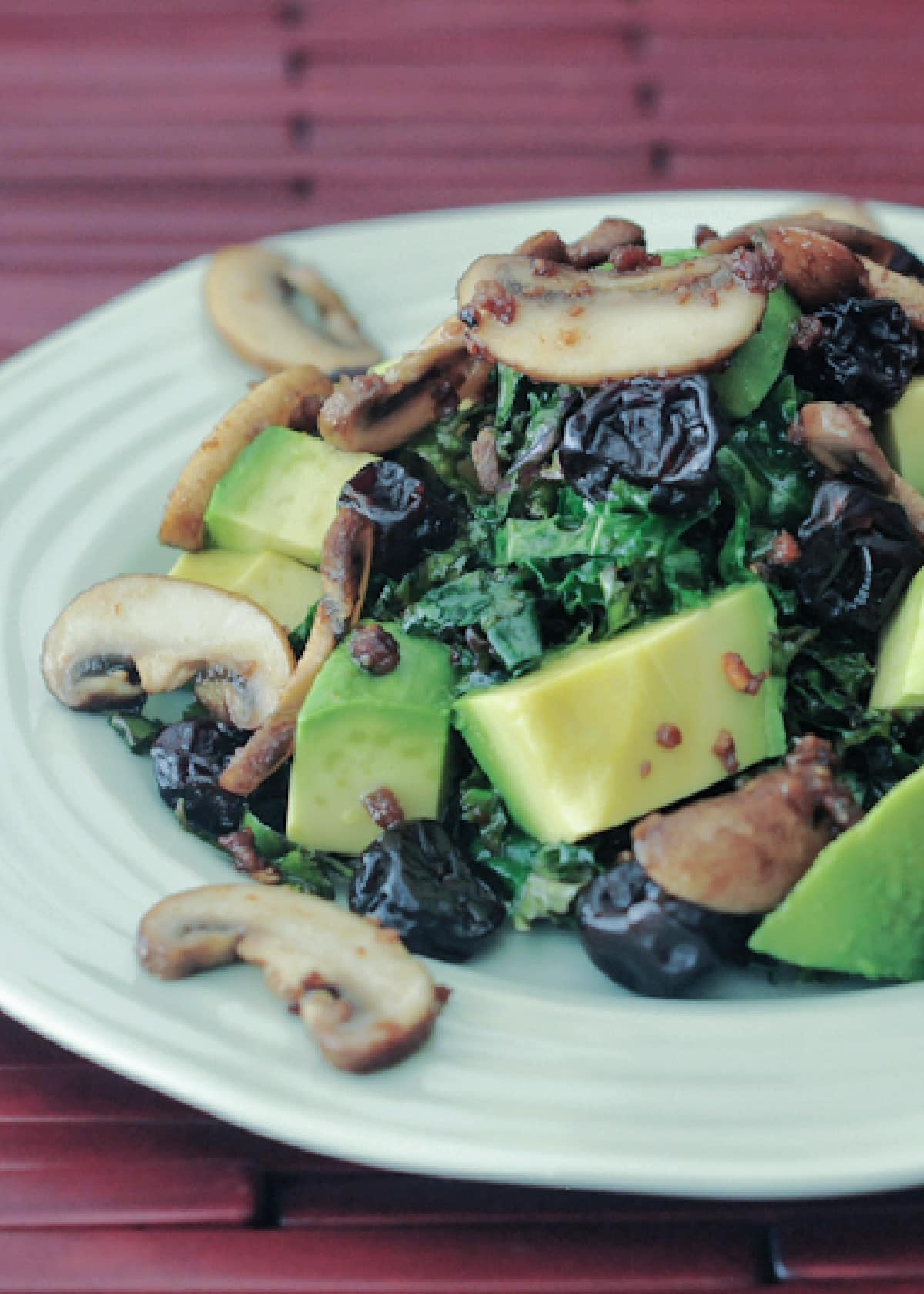 kale salad with warm bacon dressing is kale, cubed avocado, bacon flavored mushrooms on a light green plate with a reddish brown wooden slats placemat.