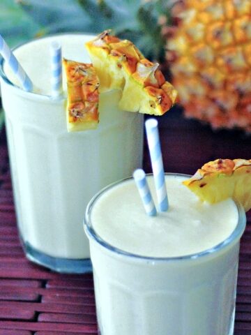 Lightened up pina coladas in glasses garnished with a wedge of pineapple and blue and white striped straws, with a whole fresh pineapple in background