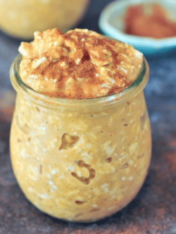 maple pumpkin overnight oats in a small glass jar on a dark grey marble tabletop, with cinnamon in a small shallow bowl and spilled on the table.