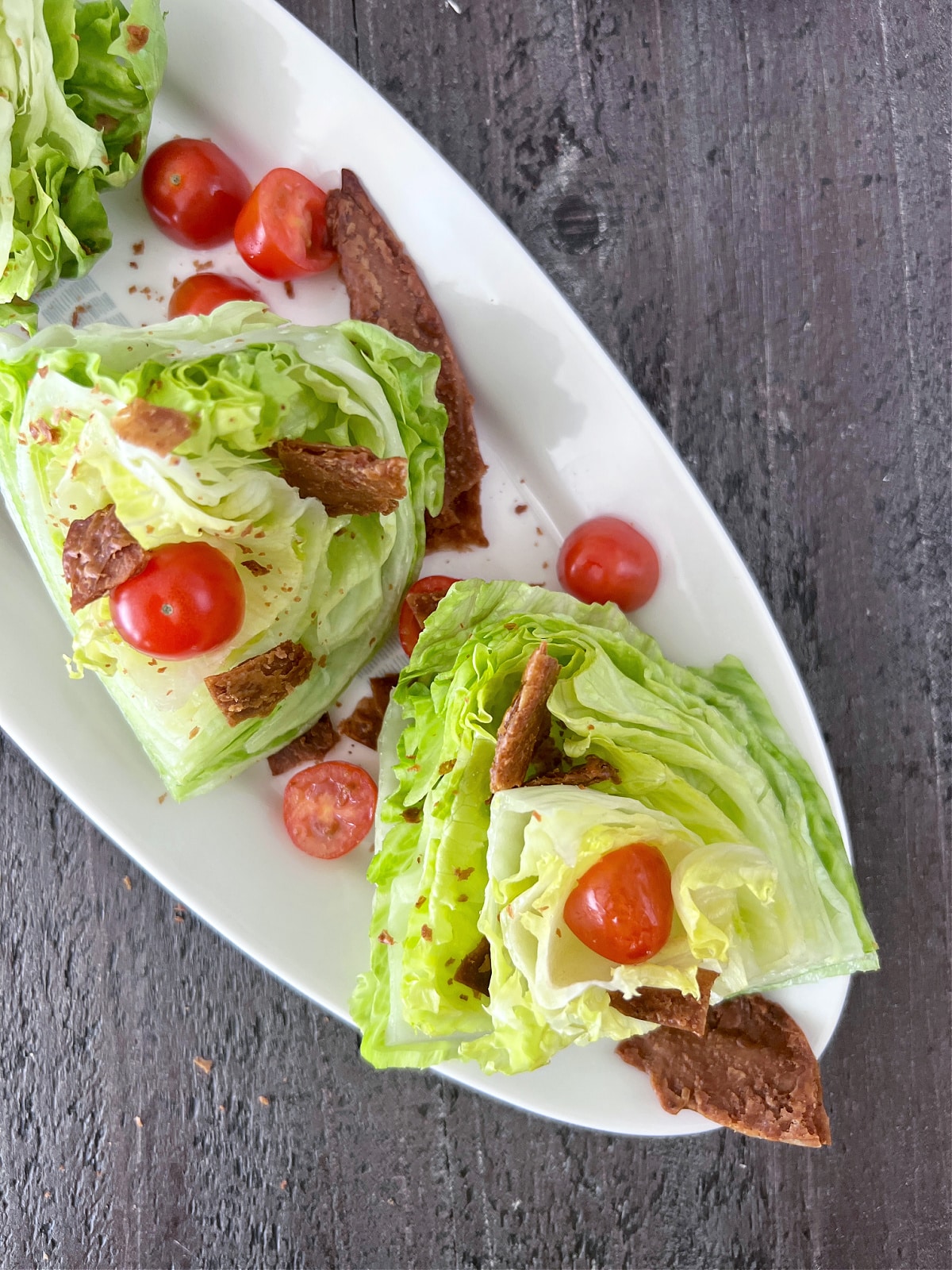 overhead view of mini wedge salads on a long white oval platter, dressed with sliced grape tomatoes, vegan bacon pieces, and chipotle ranch dressing.