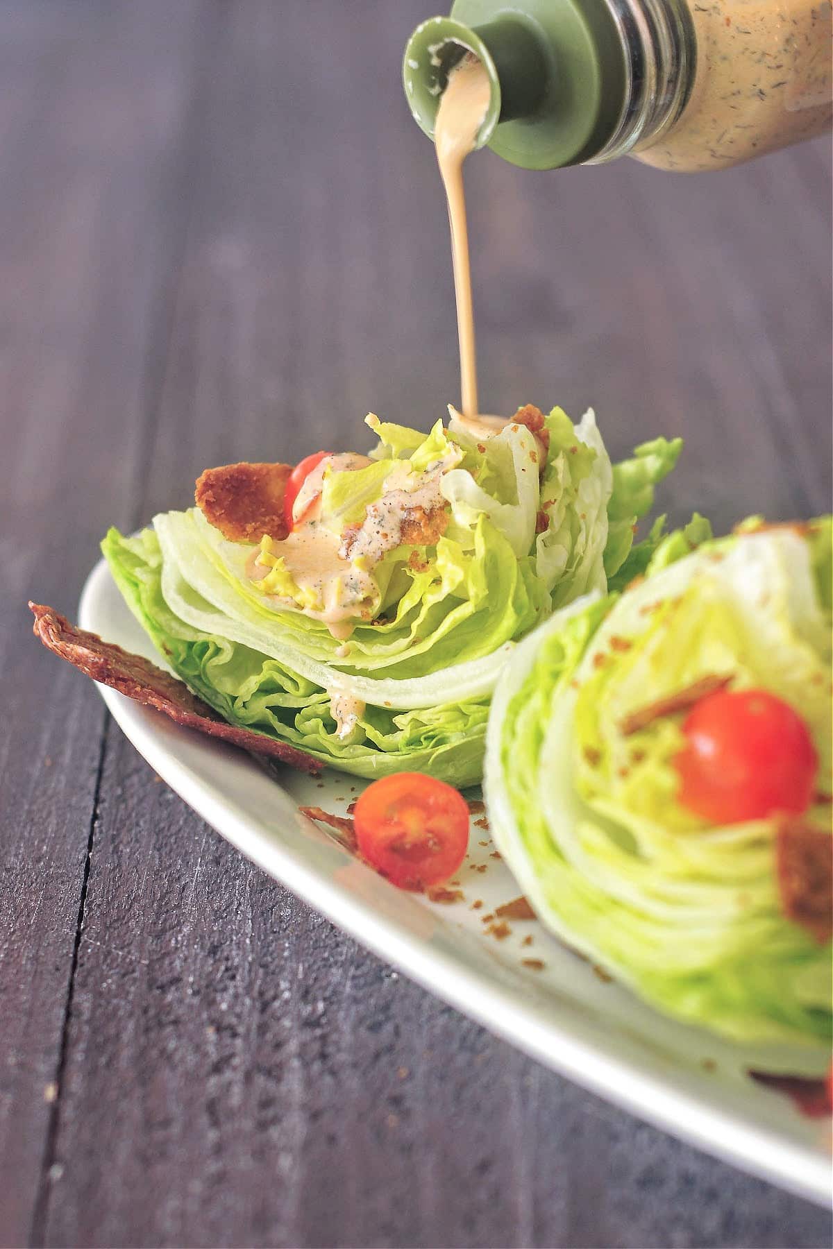 chipotle ranch dressing being poured over one mini wedge salad on a long white platter, one large piece of vegan bacon underneath the wedge, sliced grape tomato scattered on the wedge and plate.