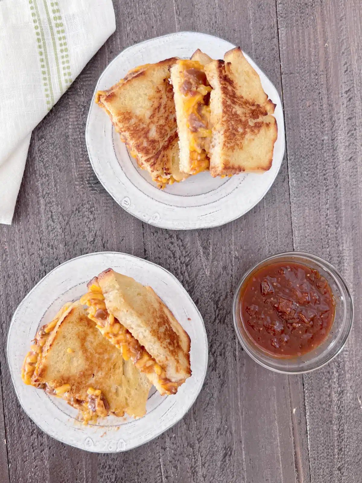 overhead view of two salsa cheddar grilled cheese sandwiches on rustic white plates next to a small glass bowl of extra salsa for dipping.