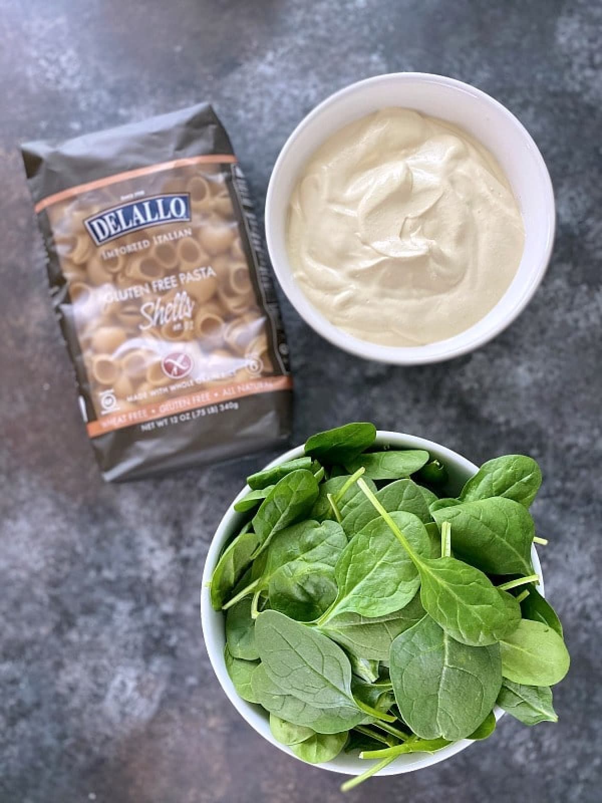 Spinach mac and cheese ingredients: a bowl of fresh spinach, a bowl of cauliflower cheese sauce, a package of gluten free pasta