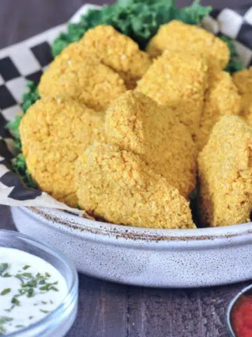 Vegan chicken nuggets in a shallow bowl with black and white checkered paper and kale garnish. dipping sauces on the side (ranch, teriyaki sauce, tomato)