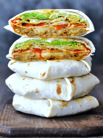 Homemade vegan crunchwrap supreme sliced in half to show filling and stacked five layers high (two halves on top of three whole crunchwraps). filling is beans, cheese, crunchy tortilla, red peppers, salsa, iceberg lettuce.