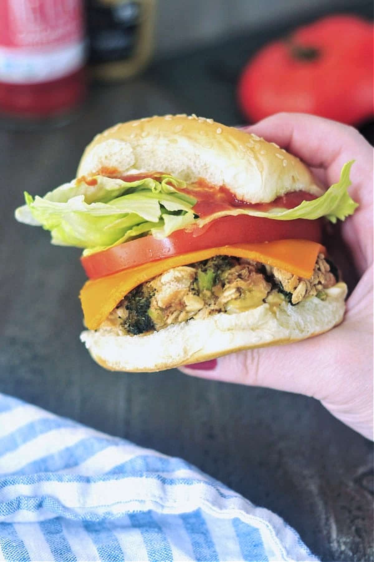 a hand holding a veggie burger dressed with cheddar, thick slice of tomato, and iceberg lettuce on a soft sesame bun