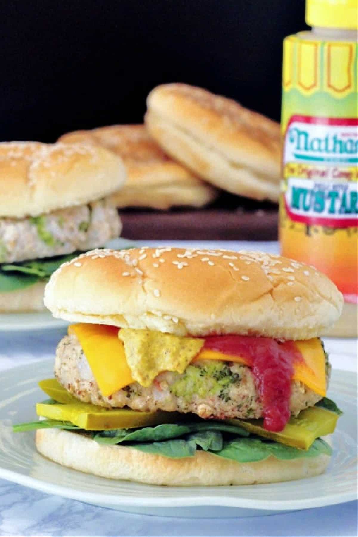 a vegan veggie burger on a plate, dressed with spinach, pickles, cheddar slice, ketchup and mustard. bottle of mustard and more burgers and buns in background, against a solid black background
