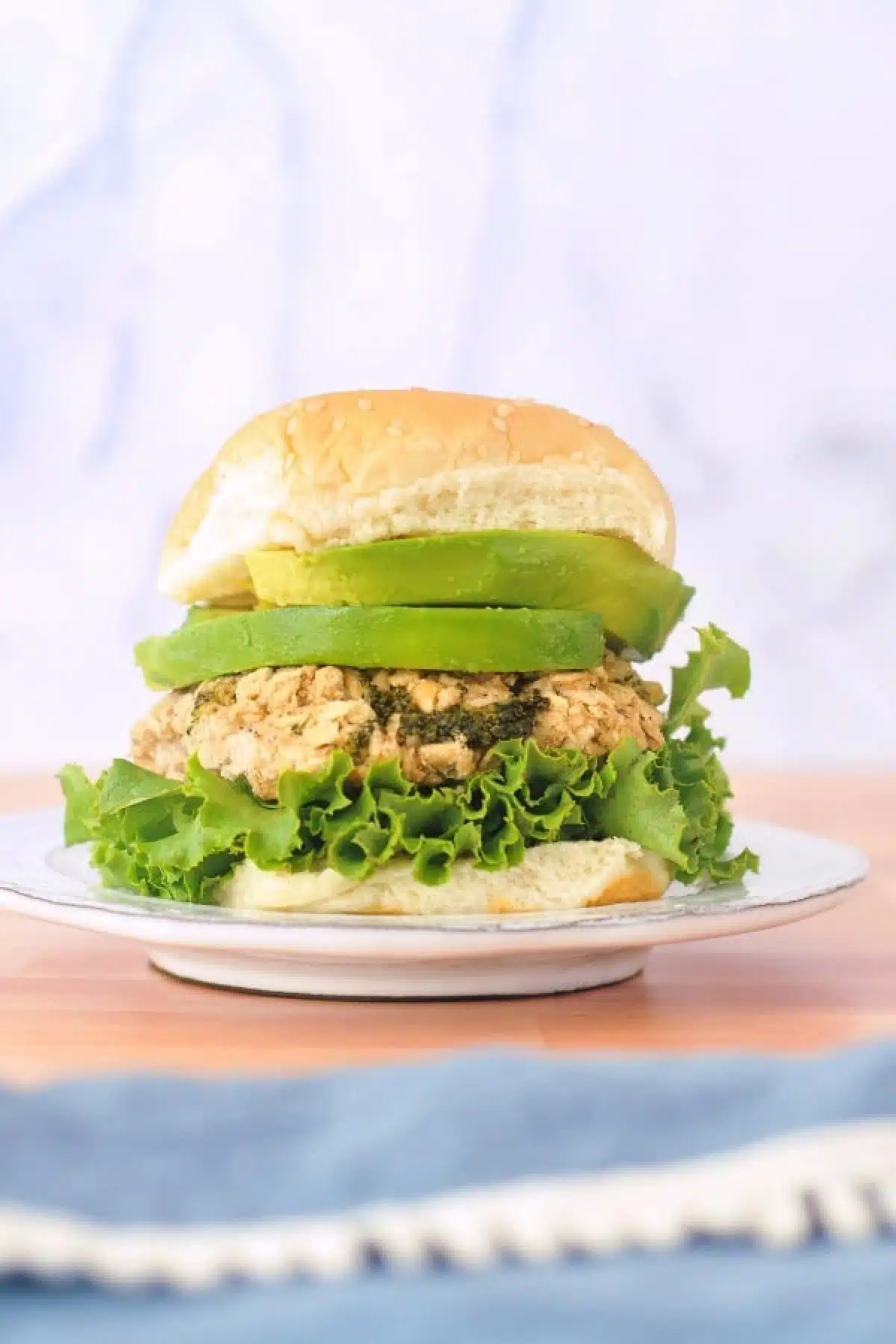 a single white bean veggie burger on a rustic white plate, dressed with curly lettuce, avocado, and a soft sesame bun