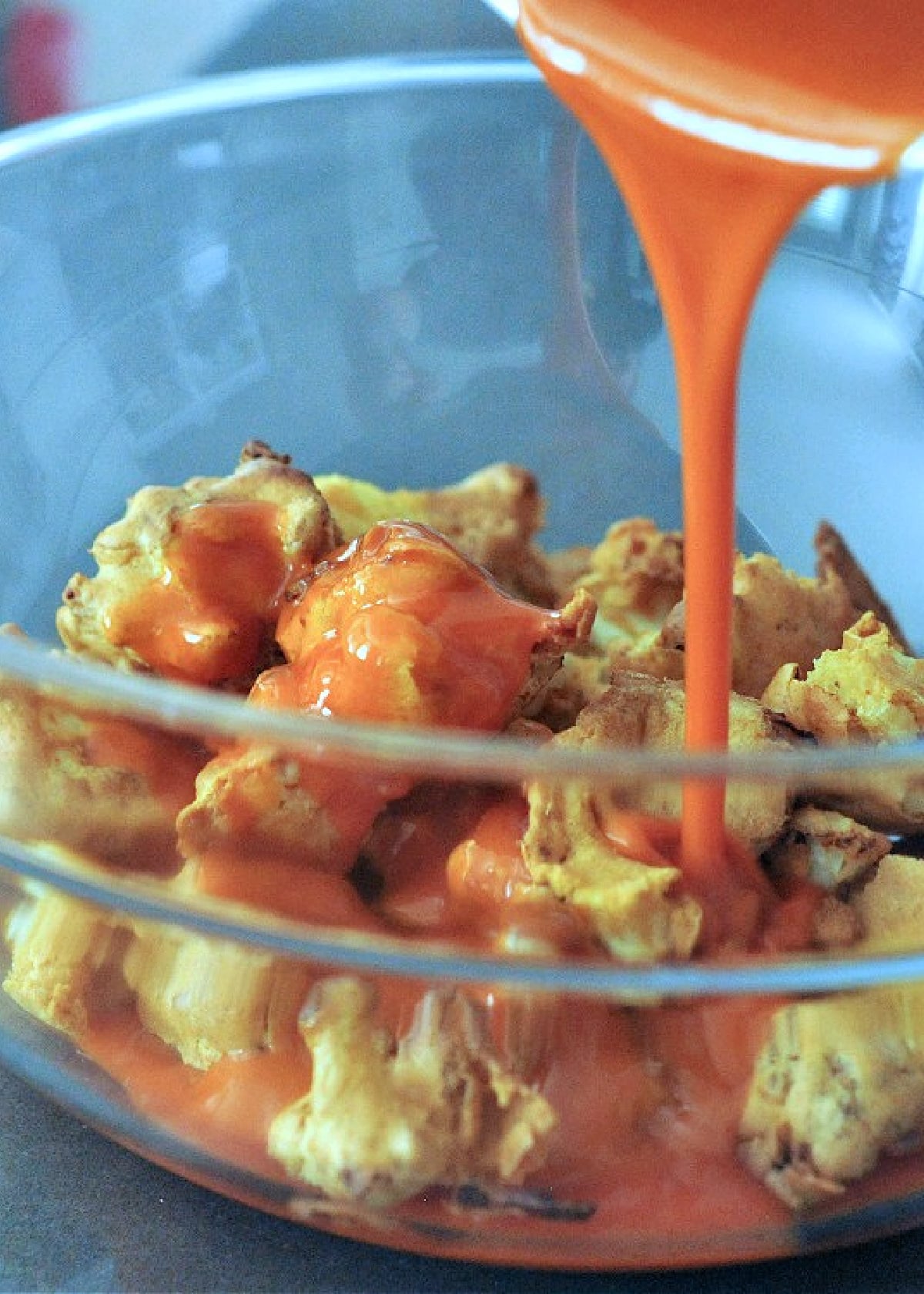 Buffalo sauce being poured over air fryer buffalo cauliflower in a clear glass bowl.