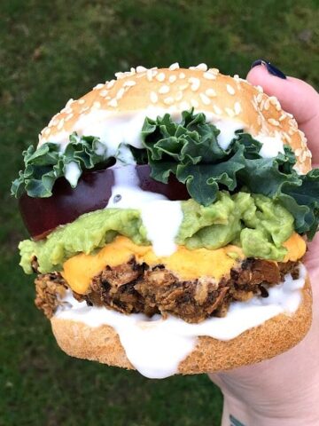 Mushroom burger held in hand, mayo dripping down side. Colorful toppings include green curly lettuce, deep red thick tomato slice, light green mashed avocado, bright yellow vegan cheese sauce, bright white vegan mayonnaise.
