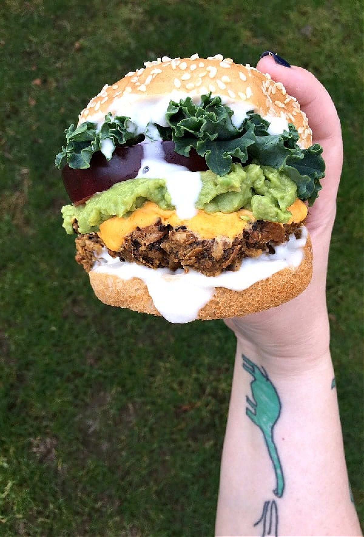 Mushroom burger held in hand, mayo dripping down side. Colorful toppings include green curly lettuce, deep red thick tomato slice, light green mashed avocado, bright yellow vegan cheese sauce, bright white vegan mayonnaise.