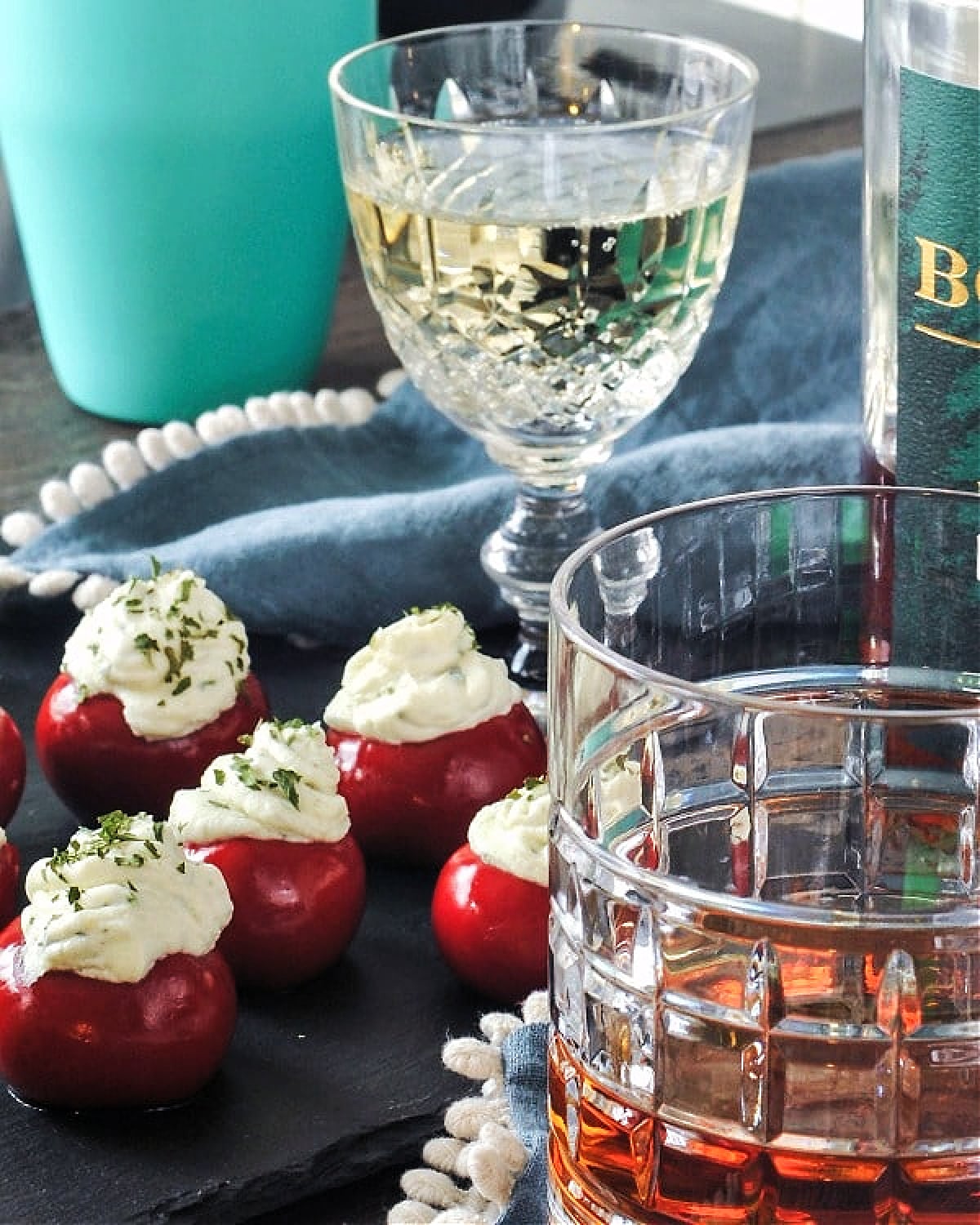 Round red peppadew sweet peppers filled with herb cheese (Boursin style) on a black slate board next to a crystal champagne glass and a rocks glass with bourbon.