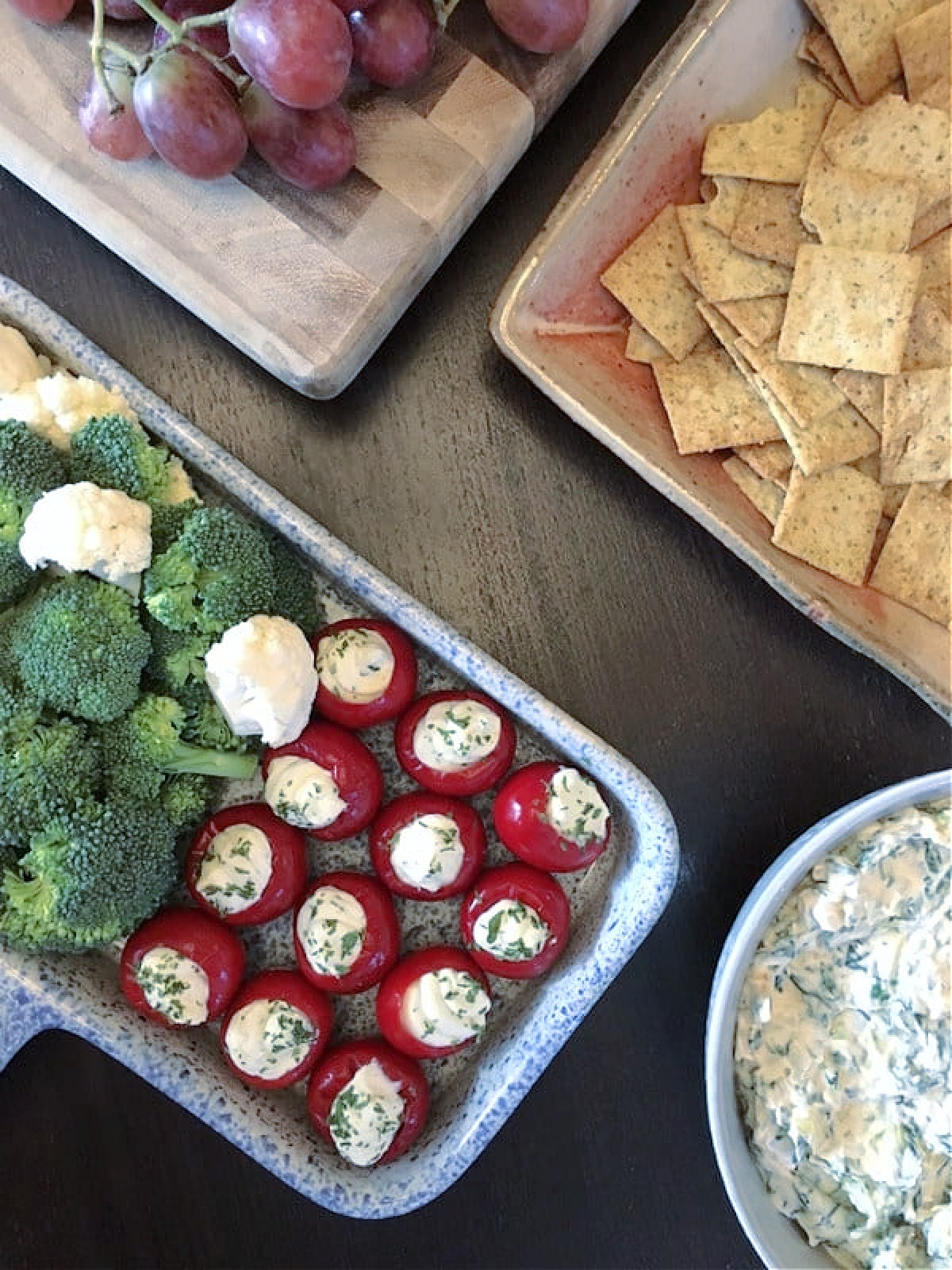Overhead view of round red peppadew sweet peppers filled with herb cheese (Boursin style) on a blue and white speckled tray next to a wood board with purple grapes, a wooden square dish with crackers, and a bowl of spinach dip.