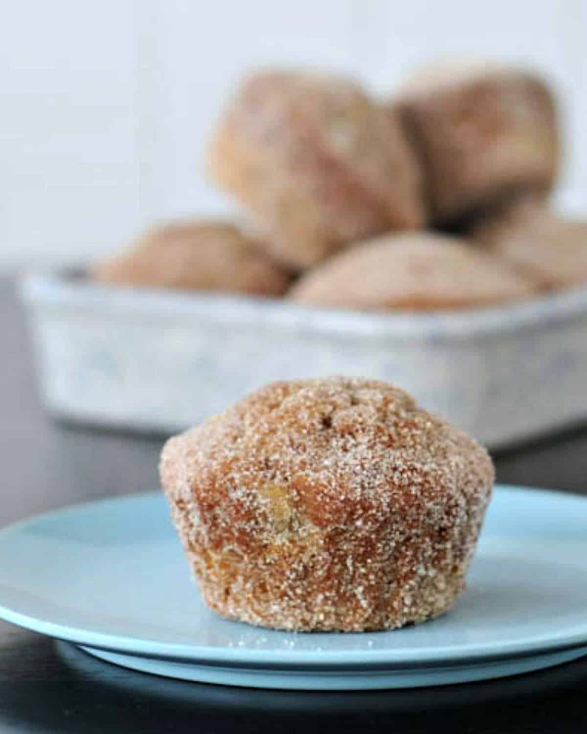 One sugar coated "donut muffin" on a light blue plate, with more muffins in a shallow walled dish blurred in background.