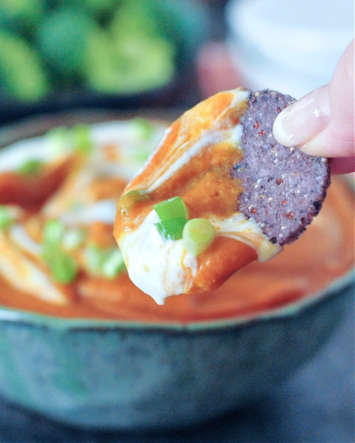 A hand holding a round blue corn chip with enchilada dip on it; a bowl of enchilada dip blurred in background.