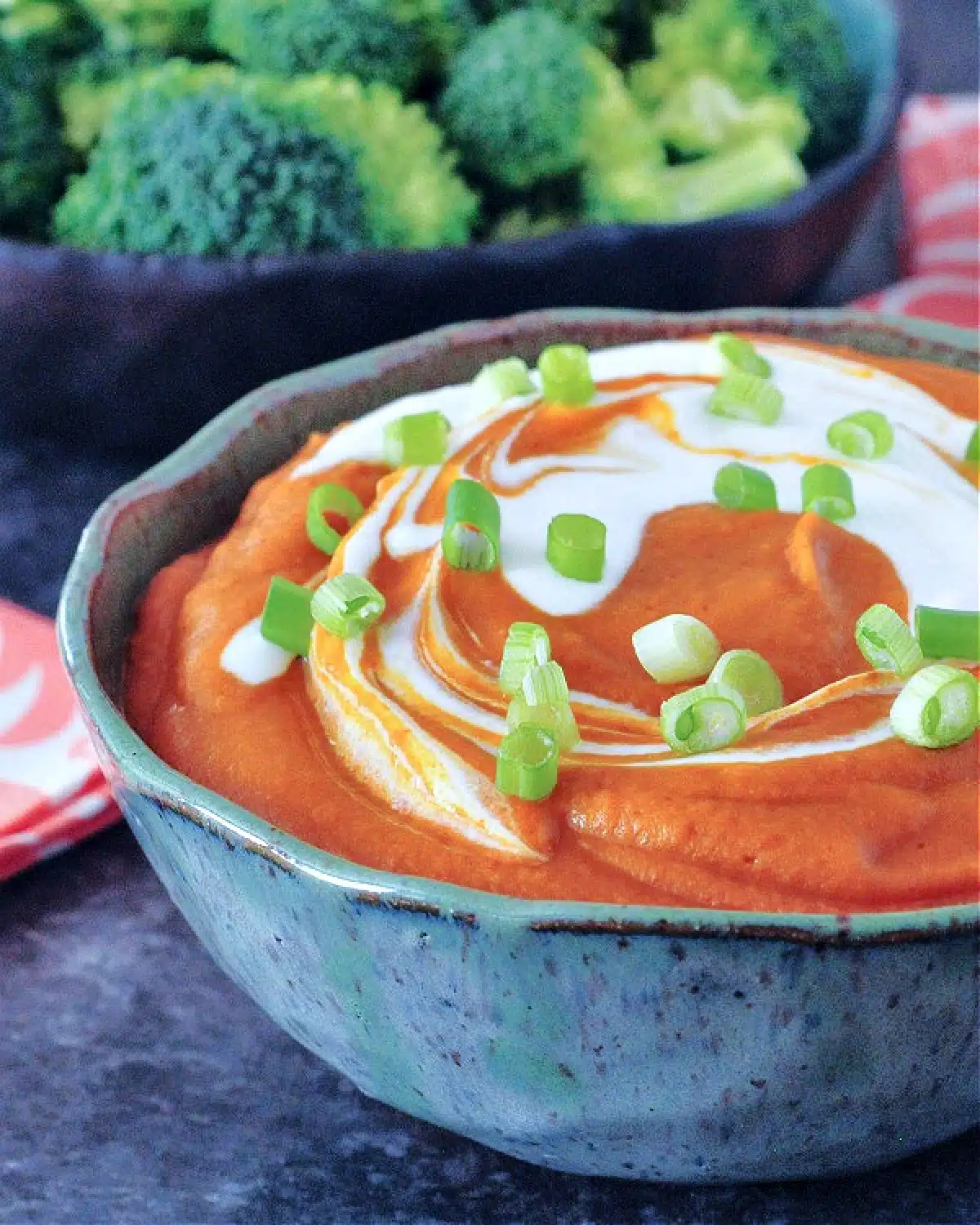 Deep orange enchilada dip with bright white sour cream swirled on top, garnished with chopped green onion. Dip is in a grey bowl with broccoli trees on the side for dipping.