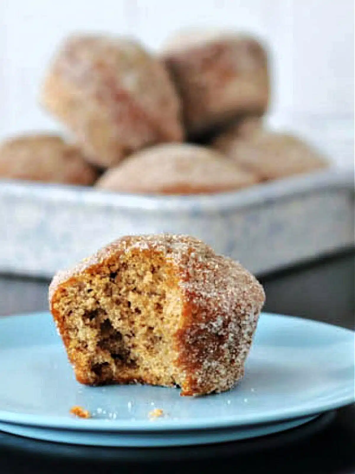 One sugar coated "donut muffin" on a light blue plate, with more muffins in a shallow walled dish blurred in background.