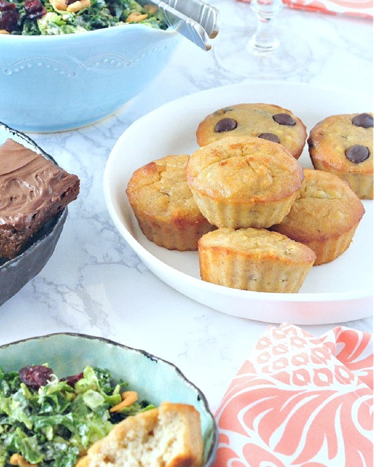 A white plate of peanut butter banana muffins among a brunch spread of salads, chocolate, and champagne.