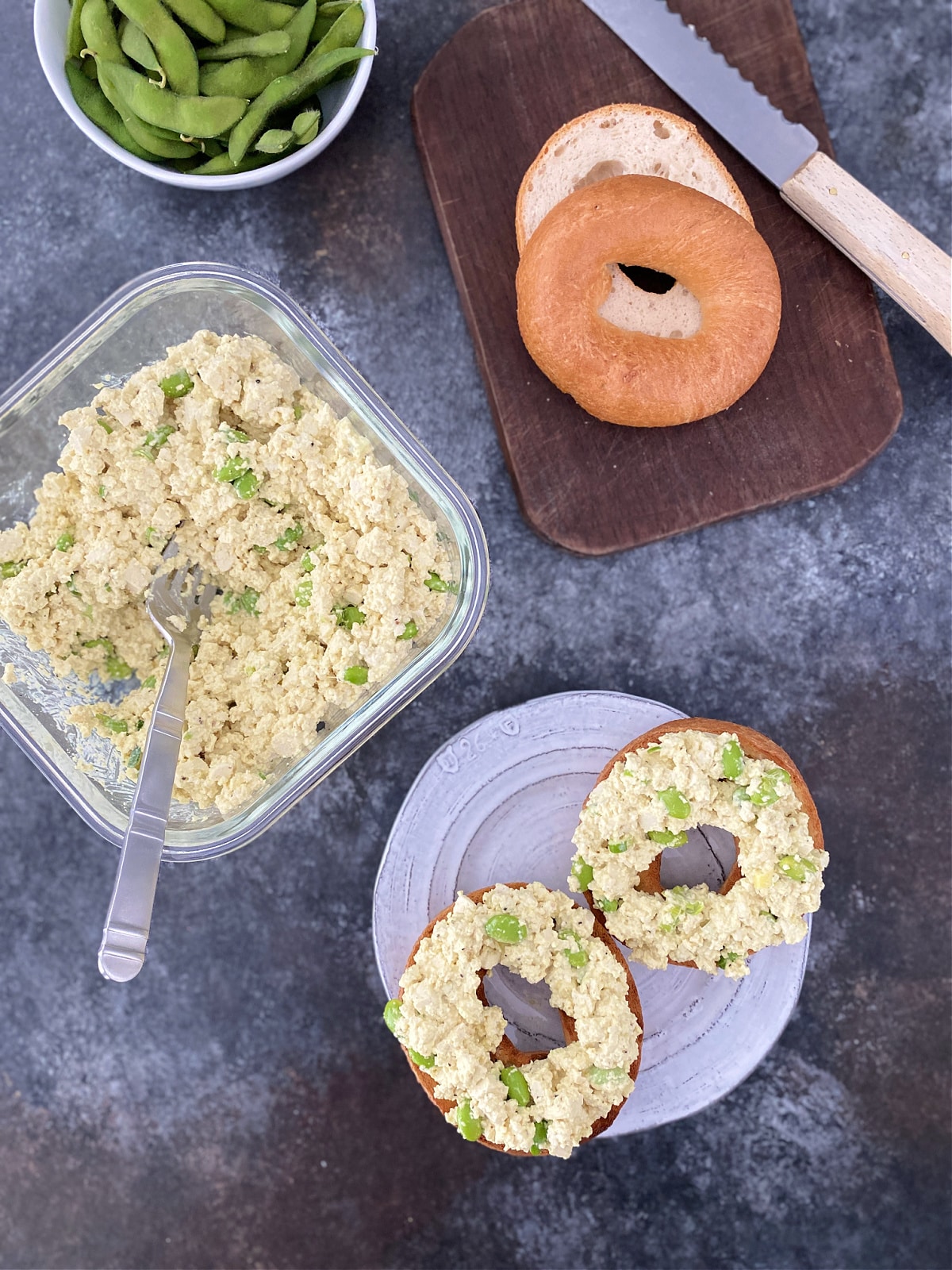 Overhead view of vegan edamame egg salad in a square glass container, with a small bowl of unshelled edamame, a small cutting board with a bagel and knife, and a plate with a toasted sliced bagel topped with egg salad.
